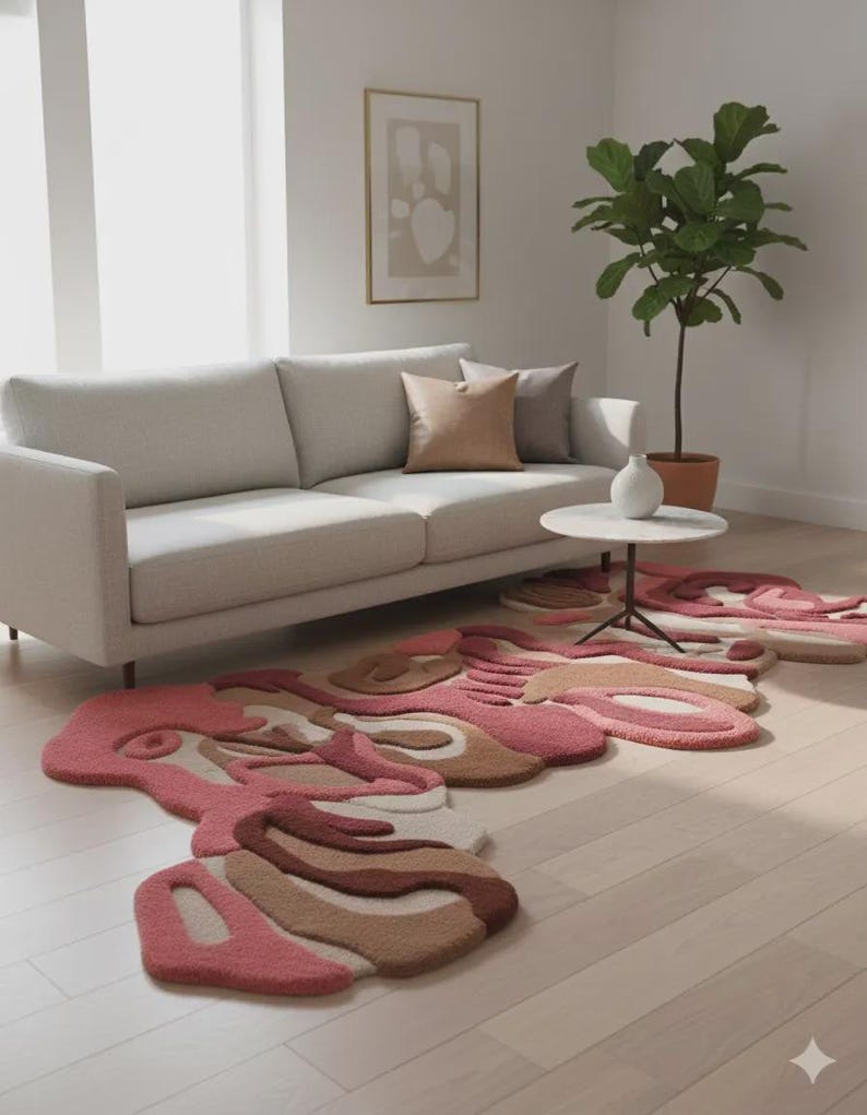 Peut inclure: Sc&egrave;ne de salon avec des tapis abstraits roses, bruns et beiges. Un canap&eacute; gris clair avec des coussins est contre un mur blanc. Une petite table ronde et une plante en pot sont &eacute;galement visibles sur le sol en bois clair.