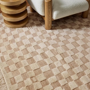 May include: A beige and cream checkered rug with a fringe border. The rug is made of a soft, textured material and has a subtle sheen.
