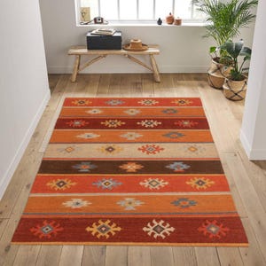 May include: A rectangular area rug with a striped pattern in shades of orange, brown, and beige. The rug features repeating geometric designs in various colors, including blue, yellow, and white. The rug is placed on a wooden floor.
