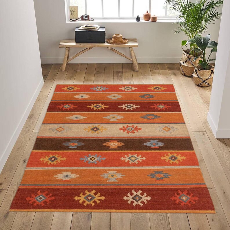 May include: A rectangular area rug with a striped pattern in shades of orange, brown, and beige. The rug features repeating geometric designs in various colors, including blue, yellow, and white. The rug is placed on a wooden floor.