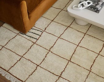 May include: A cream-colored, geometric patterned rug with brown grid lines and a fringed edge. The rug is placed on a wooden floor, with a brown velvet sofa and white coffee table in the background. The rug's design adds a touch of warmth and texture to the room.