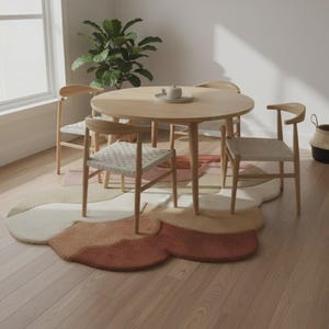 Puede incluir: Una mesa de comedor redonda de madera clara con cuatro sillas a juego. Las sillas tienen asientos tejidos. Una alfombra multicolor en tonos beige, rosa y marrón está debajo de la mesa y las sillas. Una planta y una cesta están en el fondo.