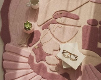 May include: A plush, abstract-shaped rug in shades of pink and mauve. The rug features a textured, raised design with a scalloped edge. A white mug, a small succulent in a pink pot, a stack of books, and eyeglasses are on the rug.