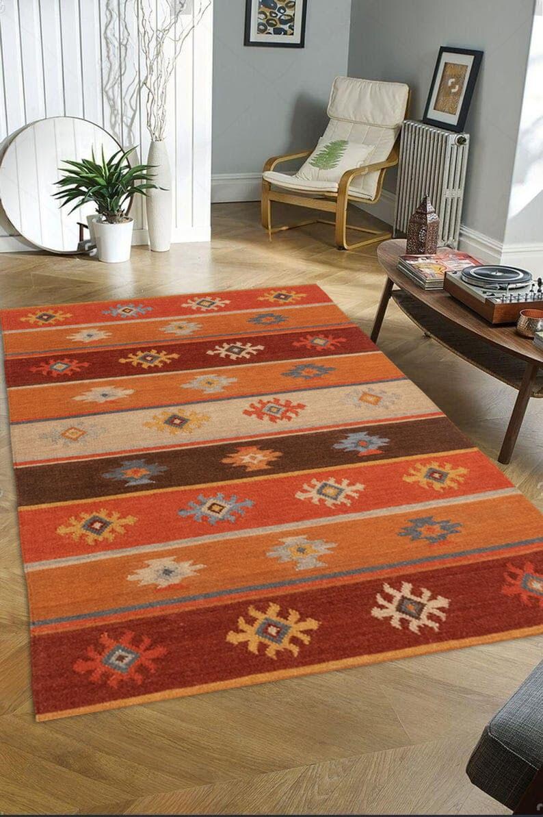 May include: Rectangular area rug with a striped pattern in shades of orange, brown, and beige. The rug features repeating geometric designs in yellow, blue, and white. The rug is placed on a wooden floor.
