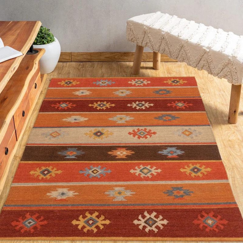 May include: A rectangular area rug with a Southwestern design. The rug features horizontal stripes in shades of orange, brown, tan, and blue. Geometric patterns are interspersed throughout the stripes. The rug is placed on a hardwood floor.
