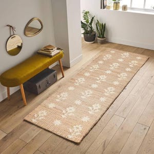 May include: A beige and white floral patterned jute rug runner. The rug is laid on a wooden floor in a hallway with a yellow bench and a potted plant.