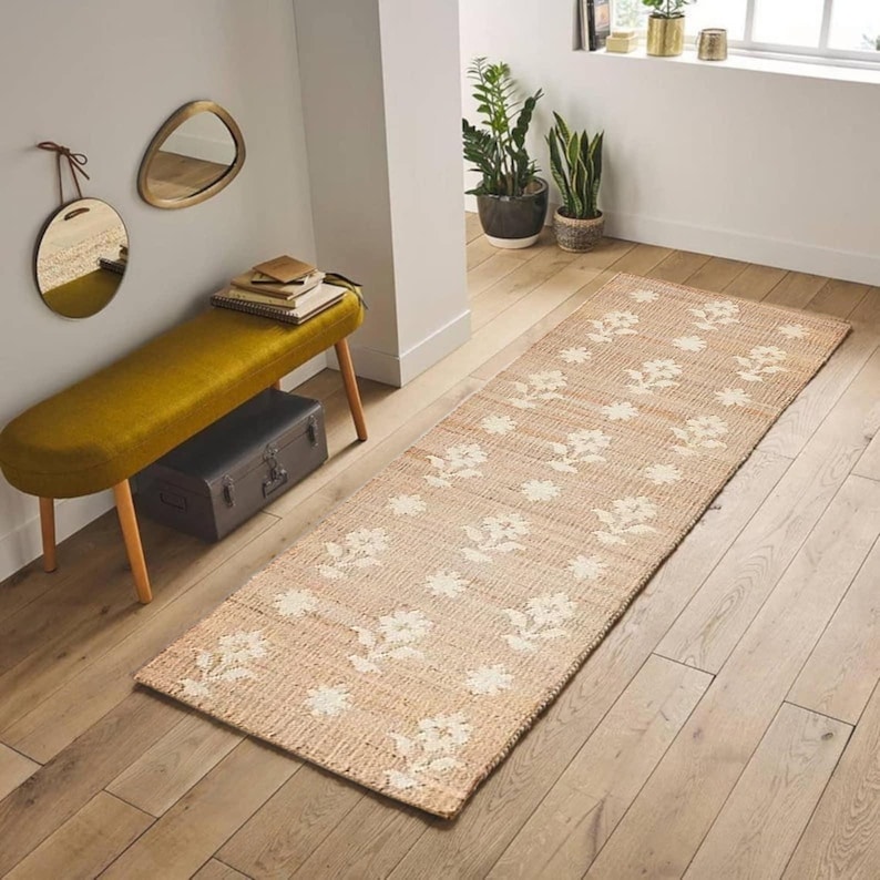 May include: A beige and white floral patterned jute rug runner. The rug is laid on a wooden floor in a hallway with a yellow bench and a potted plant.
