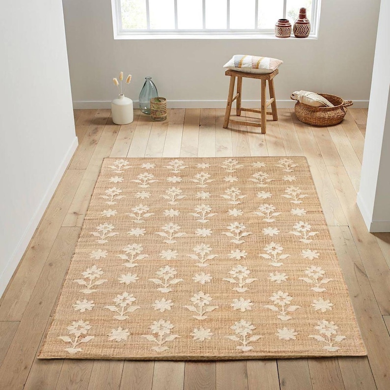 May include: A natural fiber rug with a beige background and a white floral pattern. The rug is made of jute and has a woven texture.