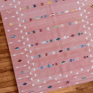 Op de afbeelding: Een roze geweven vloerkleed met een geometrisch patroon. Het vloerkleed heeft witte, blauwe, gele en rode accenten. Het vloerkleed is rechthoekig en lijkt van een zacht materiaal te zijn. Het vloerkleed ligt op een houten vloer.