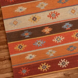 May include: A rectangular area rug with a geometric pattern. The rug features horizontal stripes in shades of orange, brown, and red, with repeating floral-like motifs in various colors. The rug is placed on a wooden floor.