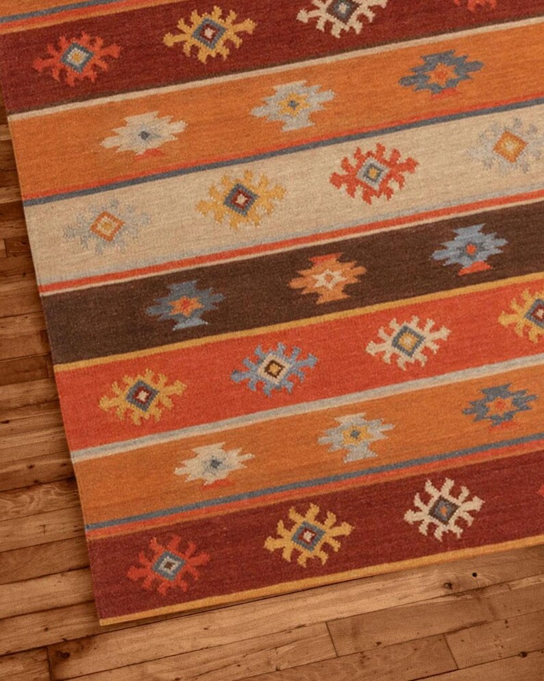May include: A rectangular area rug with a geometric pattern. The rug features horizontal stripes in shades of orange, brown, and red, with repeating floral-like motifs in various colors. The rug is placed on a wooden floor.