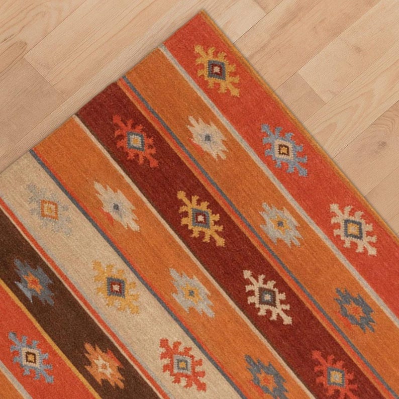 May include: A corner of a colorful rug with a geometric pattern. The rug features diagonal stripes in shades of orange, red, brown, and cream, with repeating floral-like motifs in various colors. The rug is placed on a light-colored wooden floor.
