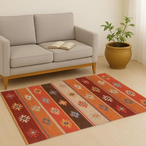 May include: A rectangular area rug with a striped pattern in shades of red, orange, brown, and beige. The rug features geometric designs and is placed on a light-colored wooden floor. A gray sofa and a potted plant are in the background.