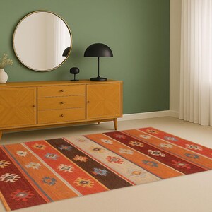 May include: A colorful area rug with a geometric pattern in shades of red, orange, brown, and cream. The rug is placed on a light-colored floor in a room with a wooden cabinet, a round mirror, and a black lamp.