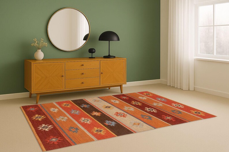 May include: A colorful area rug with a geometric pattern in shades of red, orange, brown, and cream. The rug is placed on a light-colored floor in a room with a wooden cabinet, a round mirror, and a black lamp.
