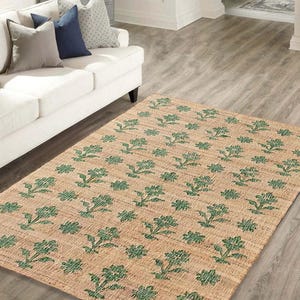 May include: A beige and green floral patterned rug with a woven texture. The rug is laid out on a hardwood floor in a living room setting.