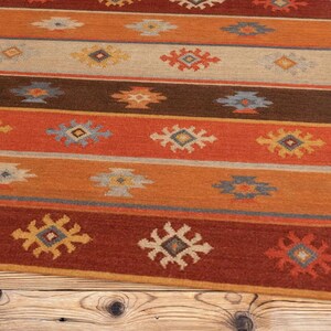 May include: A rectangular rug with a striped pattern in shades of red, orange, brown, and beige. The rug features repeating geometric designs in yellow, blue, and white. The rug is placed on a wooden surface.