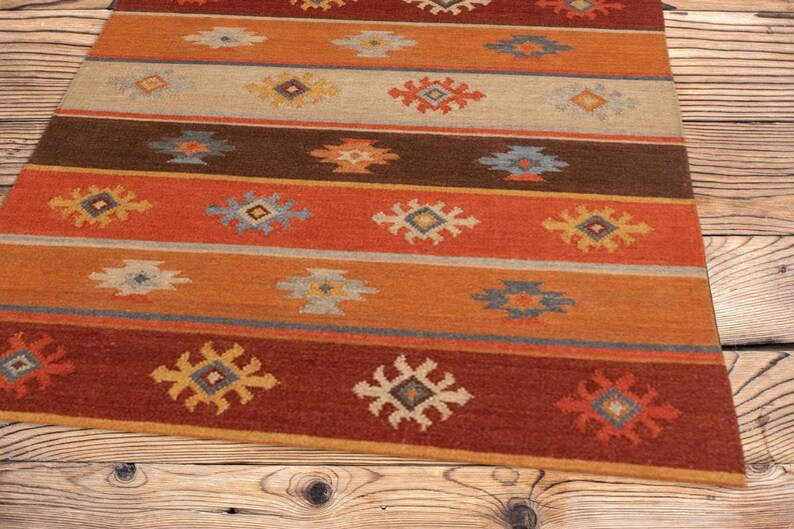 May include: A rectangular rug with a striped pattern in shades of red, orange, brown, and beige. The rug features repeating geometric designs in yellow, blue, and white. The rug is placed on a wooden surface.