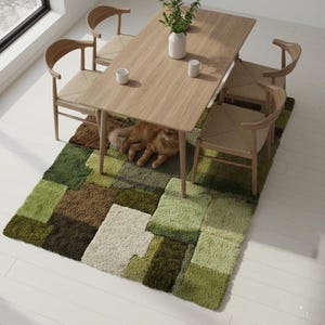 May include: A dining room scene featuring a light wood table and four wooden chairs with woven seats. A green, brown, and cream-colored geometric rug is beneath the table. A ginger cat is resting under the table. A white vase and two mugs are on the table.