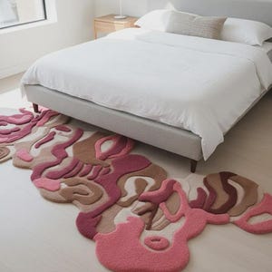 Peut inclure: Un tapis abstrait aux tons de rose, beige et marron, avec un design fluide et organique. Le tapis est pos&eacute; sur un sol en bois clair devant un lit avec une literie blanche. Le design du tapis &eacute;voque l'art moderne.
