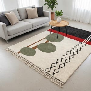 Puede incluir: Una alfombra de área color crema con un diseño geométrico. La alfombra presenta un patrón de círculos verdes, líneas marrones y formas de diamantes negros. Una sección roja y negra está en la parte superior, con rayas blancas. La alfombra tiene un borde con flecos.