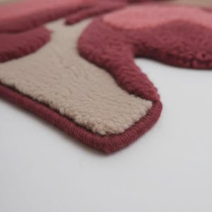 Peut inclure: Gros plan sur un tapis fait &agrave; la main avec un motif floral. Le tapis pr&eacute;sente des nuances de rose, de bordeaux et de beige. Les bords sont finis avec une bordure bordeaux, cr&eacute;ant une forme d&eacute;finie. Le tapis a un aspect doux et textur&eacute;.