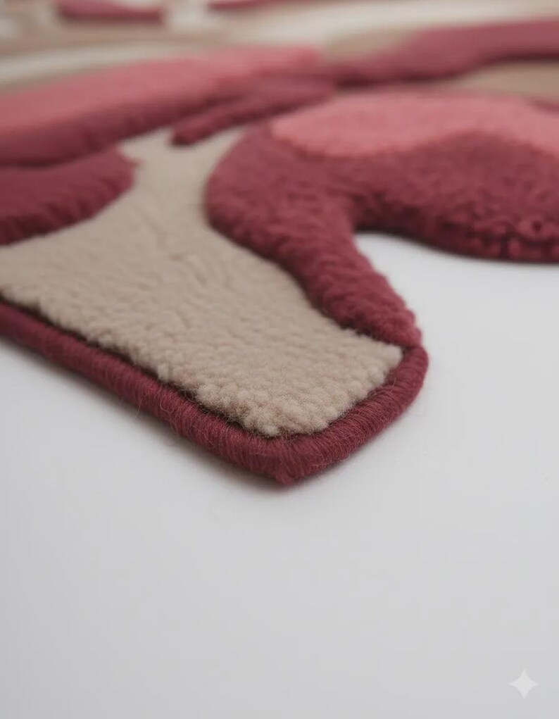 Peut inclure: Gros plan sur un tapis fait &agrave; la main avec un motif floral. Le tapis pr&eacute;sente des nuances de rose, de bordeaux et de beige. Les bords sont finis avec une bordure bordeaux, cr&eacute;ant une forme d&eacute;finie. Le tapis a un aspect doux et textur&eacute;.