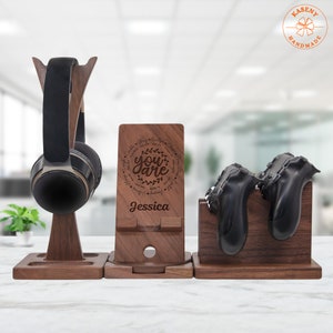 May include: Wooden stand with three compartments for organizing gaming accessories. The stand has a headphone holder, a phone stand with a personalized message, and a holder for two game controllers. The phone stand has the text "You are" surrounded by positive words and the name "Jessica".