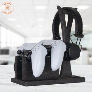 May include: A dark stained wooden gaming station holds two white game controllers and a black headset. The station has a slot for small items and a logo in the upper left corner.