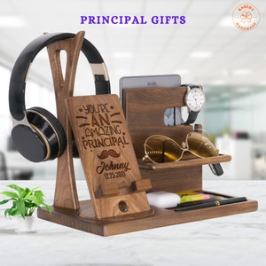 May include: Wooden desk organizer with a personalized message that reads "You're an amazing principal. Johnny 12.25.2021". The organizer has slots for a phone, watch, glasses, and pens. It also has a stand for headphones and a small shelf for a book or other items.