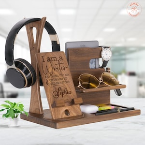 May include: A wooden desk organizer with a headphone stand, phone holder, and compartments for accessories. The organizer has the text "I am a Writer" and includes a watch, sunglasses, and a Kindle. A small plant is on the left.