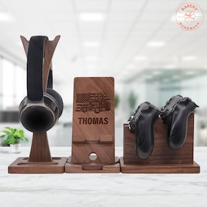 May include: A wooden desk organizer set featuring a headphone stand, a phone stand with the name "THOMAS" and a truck design, and a stand for two game controllers. The headphone stand holds black headphones. The set is made of dark wood.
