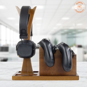May include: A wooden headphone and controller stand. The stand is made of dark brown wood and features a tall, angled headphone holder and a separate section for two game controllers. A pair of black headphones and two black controllers are displayed.
