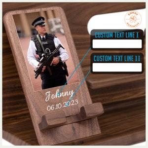 May include: A wooden phone stand with a photo of a police officer in uniform holding a gun. The stand has a slot for a phone and a space for a personalized message. The message reads "Johnny 06.10.2023".