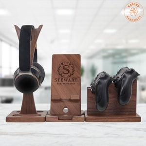 May include: A wooden stand with three sections for organizing gaming accessories. The left section has a hook for headphones, the middle section has a phone stand with a cutout for charging, and the right section has two slots for controllers. The middle section is engraved with a wreath, the letter "S", and the text "STEWART EST. 06.10.2023".
