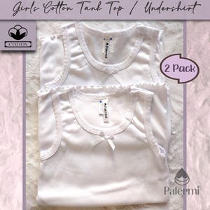 May include: Two pack of white cotton tank tops with lace trim. The tank tops have a bow on the front and a "Palermi" label on the side. The image includes the text "Girls Cotton Tank Top / Undershirt" and "2 Pack".