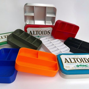Könnte beinhalten: Eine Sammlung von sechs verschiedenen farbigen Altoids-Dosen mit verschiedenen Fächern im Inneren. Die Dosen sind aus Metall und haben eine Vielzahl von Farben, darunter Blau, Orange, Grün, Rot, Weiß und Silber. Die Dosen sind alle leer und bereit, mit kleinen Gegenständen gefüllt zu werden.