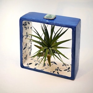Op de afbeelding: Een vierkante, blauw geschilderde houten plantenbak met een decoratief bloemenpatroon aan de binnenkant. De doos bevat een groene luchtplant. Een kleine, rechthoekige metalen accent is bevestigd aan de bovenkant van de doos.