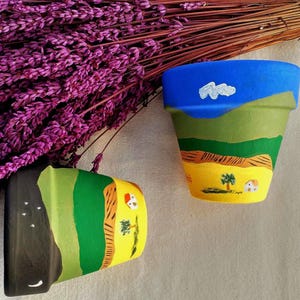 Peut inclure: Deux pots de fleurs en terre cuite peints à la main avec des paysages. L'un représente une scène de jour avec un ciel bleu, des collines vertes et un champ jaune avec un arbre et une maison. L'autre pot montre une scène de nuit.