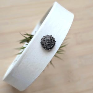 May include: A white ceramic ring-shaped planter with a smooth matte finish. Attached to the outer surface is a small, round, dark metallic decorative stud featuring a radial pattern of embossed lines and tiny circular beads along the edge.