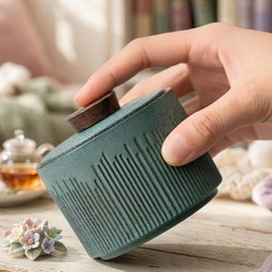 May include: A teal ceramic tea caddy with a wooden lid. The caddy has a textured, vertical line design. The lid is a small, round piece of dark wood. The caddy is held in a hand.