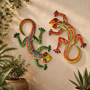 May include: Two metal gecko wall sculptures featuring vibrant, multi-coloured patterns in shades of red, yellow, green, and blue. Each lizard has a curved tail, four splayed feet, and large, prominent eyes with red and yellow circular markings.