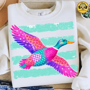 May include: A white crewneck sweatshirt featuring a graphic of a flying duck. The duck is decorated with vibrant pink, purple, and teal patterns, including leopard spots, stars, and gingham checks. Behind the duck are horizontal teal brushstrokes.