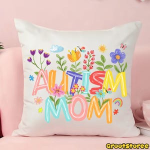 Puede incluir: Un coj&iacute;n cuadrado blanco con las palabras "AUTISM MOM" en letras coloridas y caprichosas. Las letras est&aacute;n rodeadas de una variedad de flores ilustradas, mariposas y un arco&iacute;ris. El coj&iacute;n est&aacute; sobre una superficie rosa.