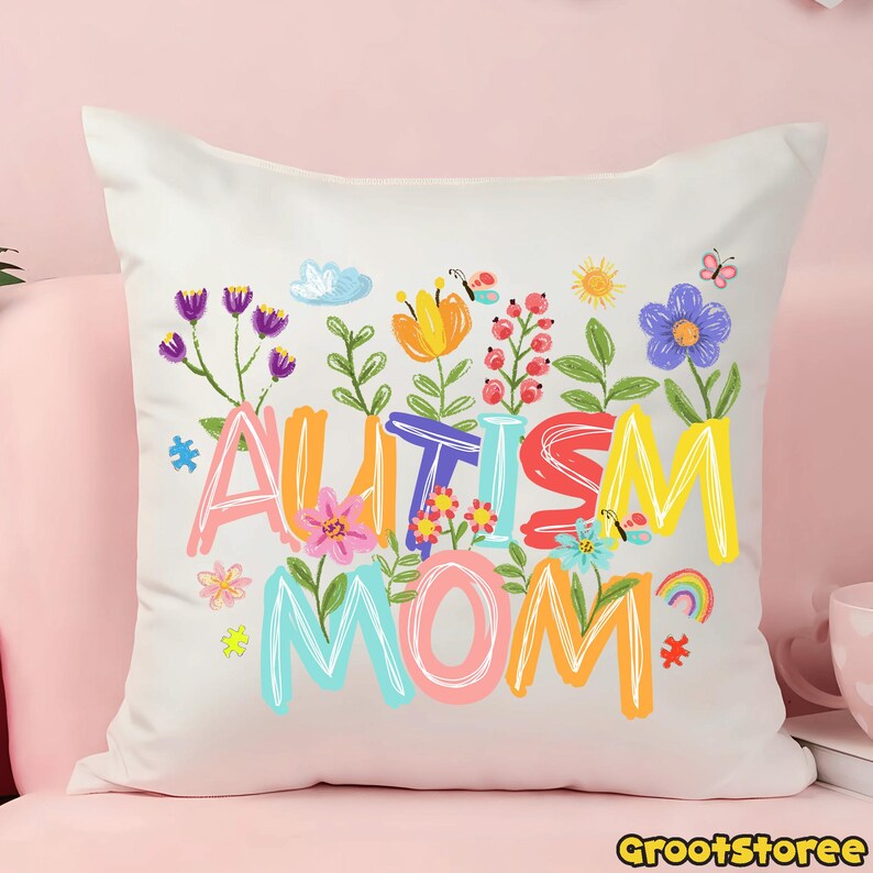 Puede incluir: Un coj&iacute;n cuadrado blanco con las palabras "AUTISM MOM" en letras coloridas y caprichosas. Las letras est&aacute;n rodeadas de una variedad de flores ilustradas, mariposas y un arco&iacute;ris. El coj&iacute;n est&aacute; sobre una superficie rosa.