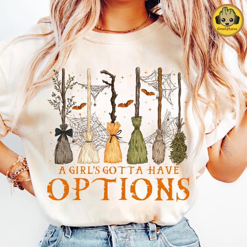 May include: Cream-colored t-shirt featuring a Halloween design. The graphic includes six different broom designs, bats, and spiderwebs. The text "A Girl's Gotta Have Options" is printed in orange below the brooms.