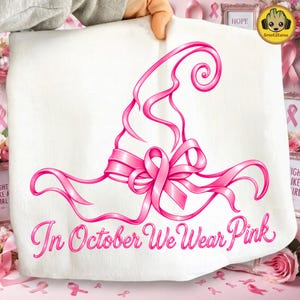 Könnte beinhalten: Ein weißer Stoffartikel mit einer rosa Grafik eines Hexenhuts, der mit einer Schleife zur Brustkrebsvorsorge verziert ist. Unter dem Hut steht der Text In October We Wear Pink in einer kursiven Schriftart in verschiedenen Rosatönen.