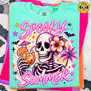 May include: A mint green t-shirt featuring a graphic of a skeleton holding a tropical cocktail with an orange slice and umbrella. The design includes a hibiscus flower, a palm tree, bats, spiders, and stars, with the text SPOOKY SUMMER in pink.