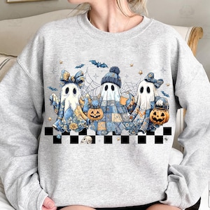 K&ouml;nnte beinhalten: Hellgraues Sweatshirt mit einer Grafik von drei Cartoon-Geistern in blau-gelben Mustern, die K&uuml;rbislaternen und Getr&auml;nke halten. Das Design umfasst Flederm&auml;use, Spinnweben und Sterne mit einem schwarz-wei&szlig;en Schachbrettmuster.