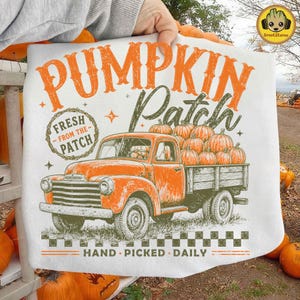 May include: A white t-shirt featuring an orange graphic of a vintage pickup truck loaded with pumpkins. The text reads PUMPKIN Patch, FRESH FROM THE PATCH, and HAND PICKED DAILY. The design includes orange and green accents with a checkered border.